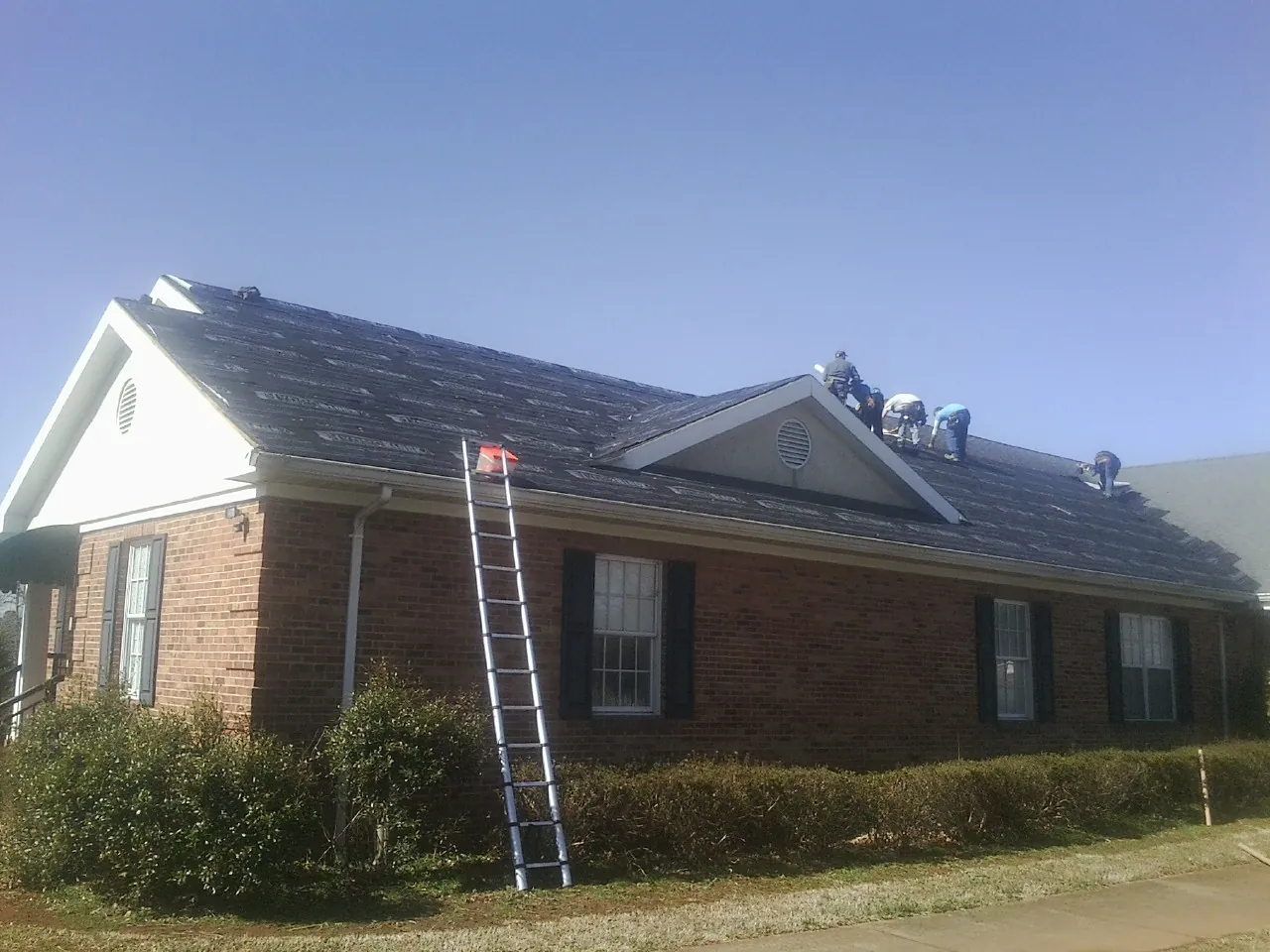 Crew tearing shingles off a brick ranch roof with a ladder set — Emergency Roof Repair work by our crew in New Riegel