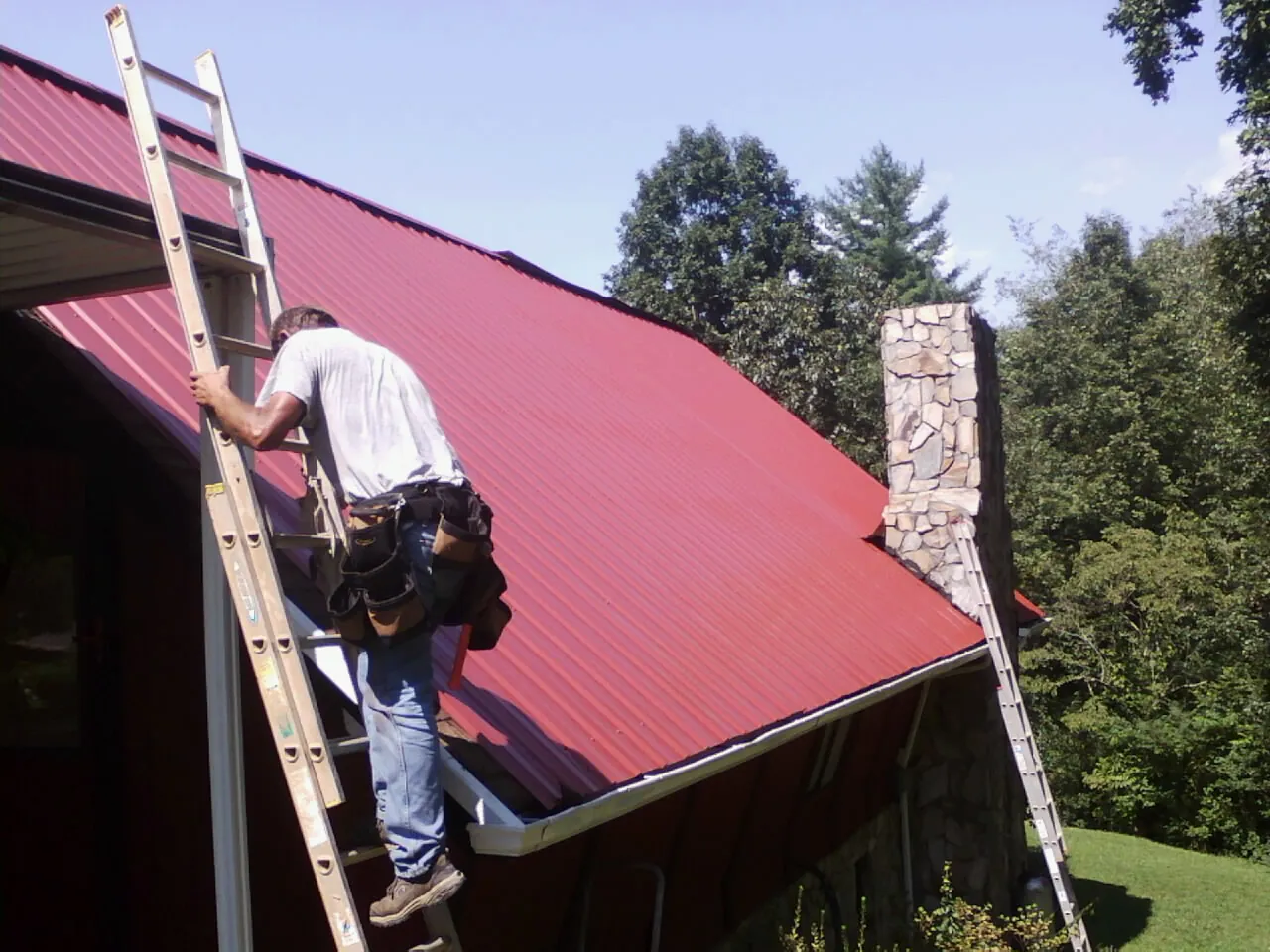 Roofer on a ladder beside a red metal roof and stone chimney — Soffit & Fascia Repair by Roofing Brothers in New Riegel